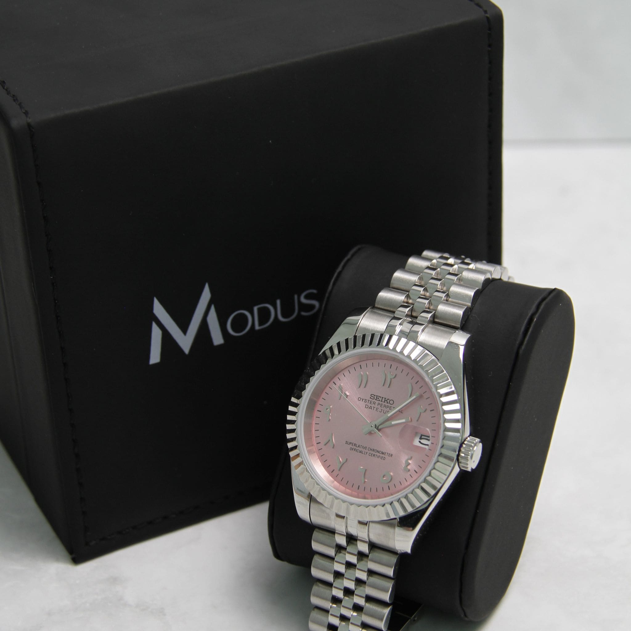 Montre Seiko Mod datejust rose sur coussin noir avec écrin en arrière plan.