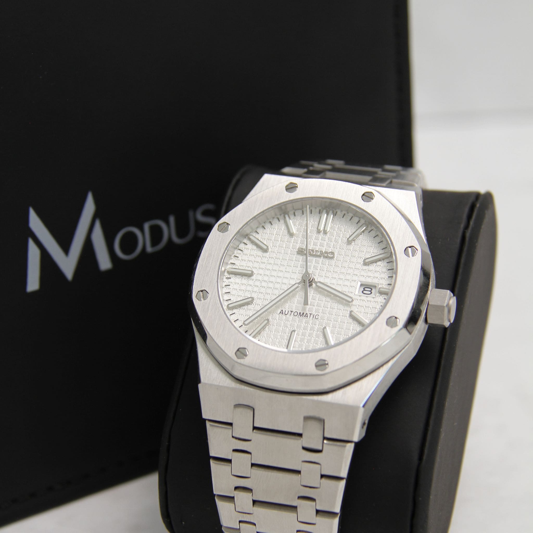 Montre Seiko Mod Royal oak blanc sur coussin noir avec écrin en arrière plan.