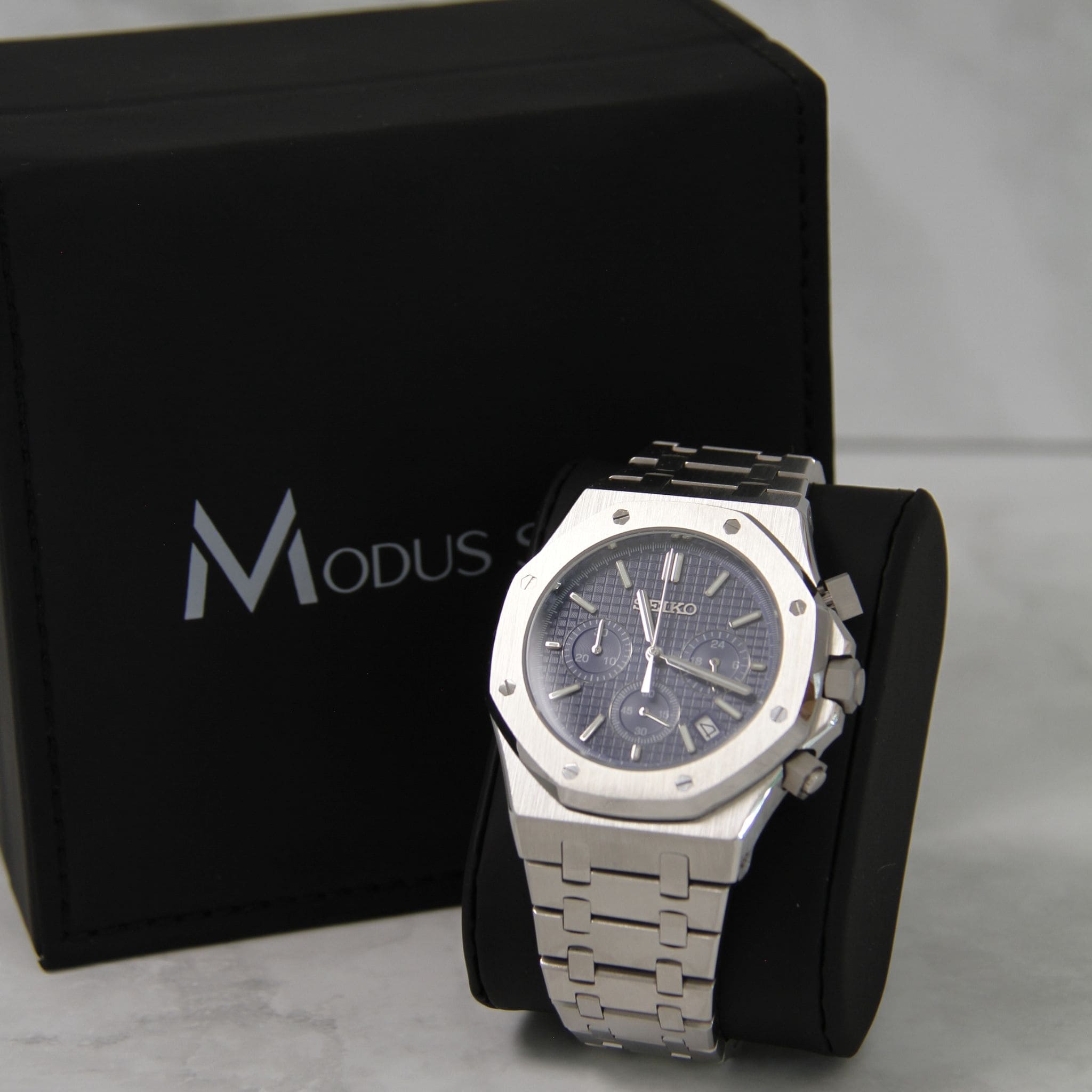 Montre Seiko Mod royal oak chronos bleu sur coussin noir avec écrin en arrière plan.