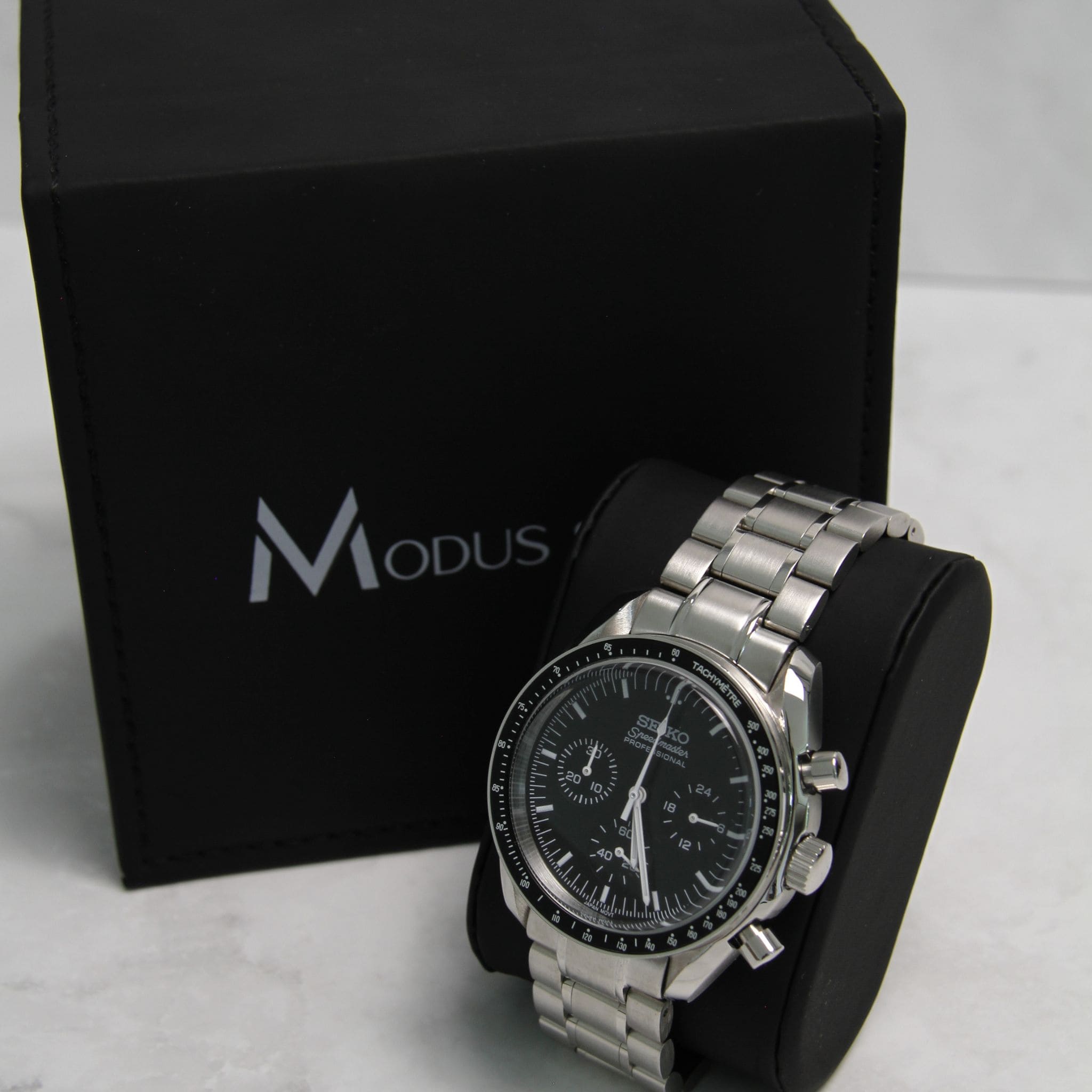 Montre Seiko Mod speedmaster noir sur coussin noir avec écrin en arrière plan