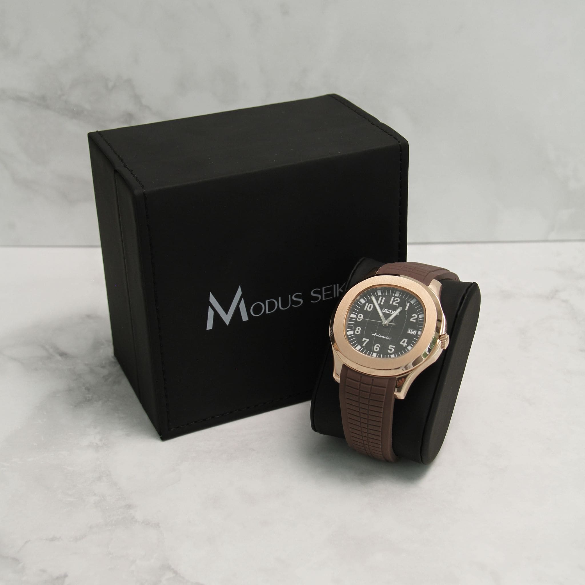 Montre Seiko Mod aquanaut or rose noir sur coussin noir avec écrin en arrière plan.