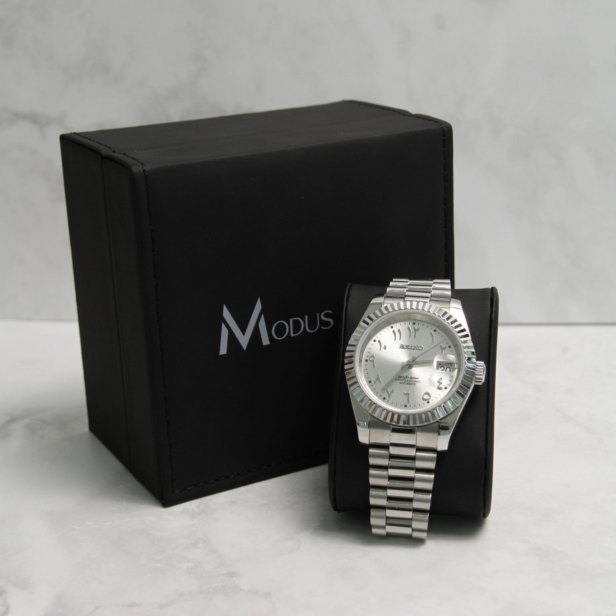 Montre Seiko Mod datejust arabic gris argenté sur coussin noir avec écrin en arrière plan.