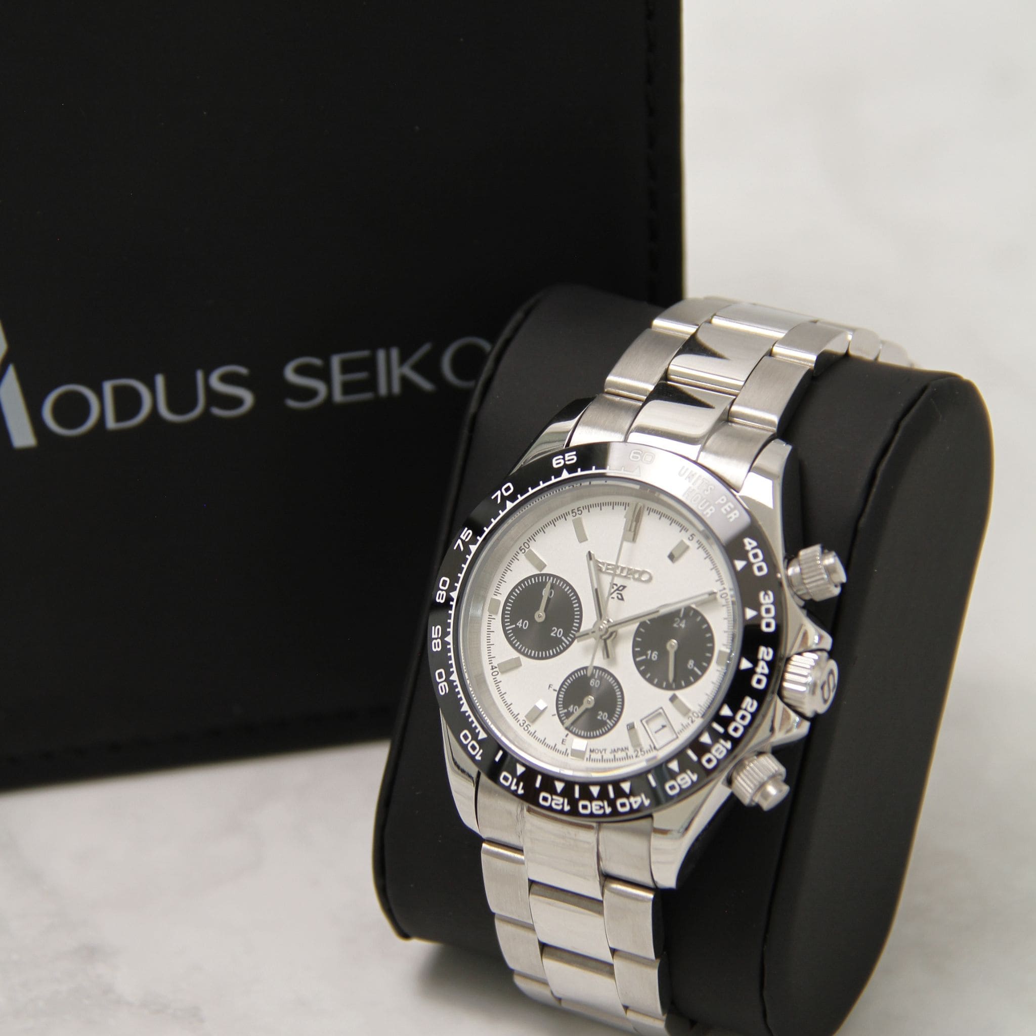 Montre Seiko Mod daytona blanc sur coussin noir avec écrin en arrière plan.
