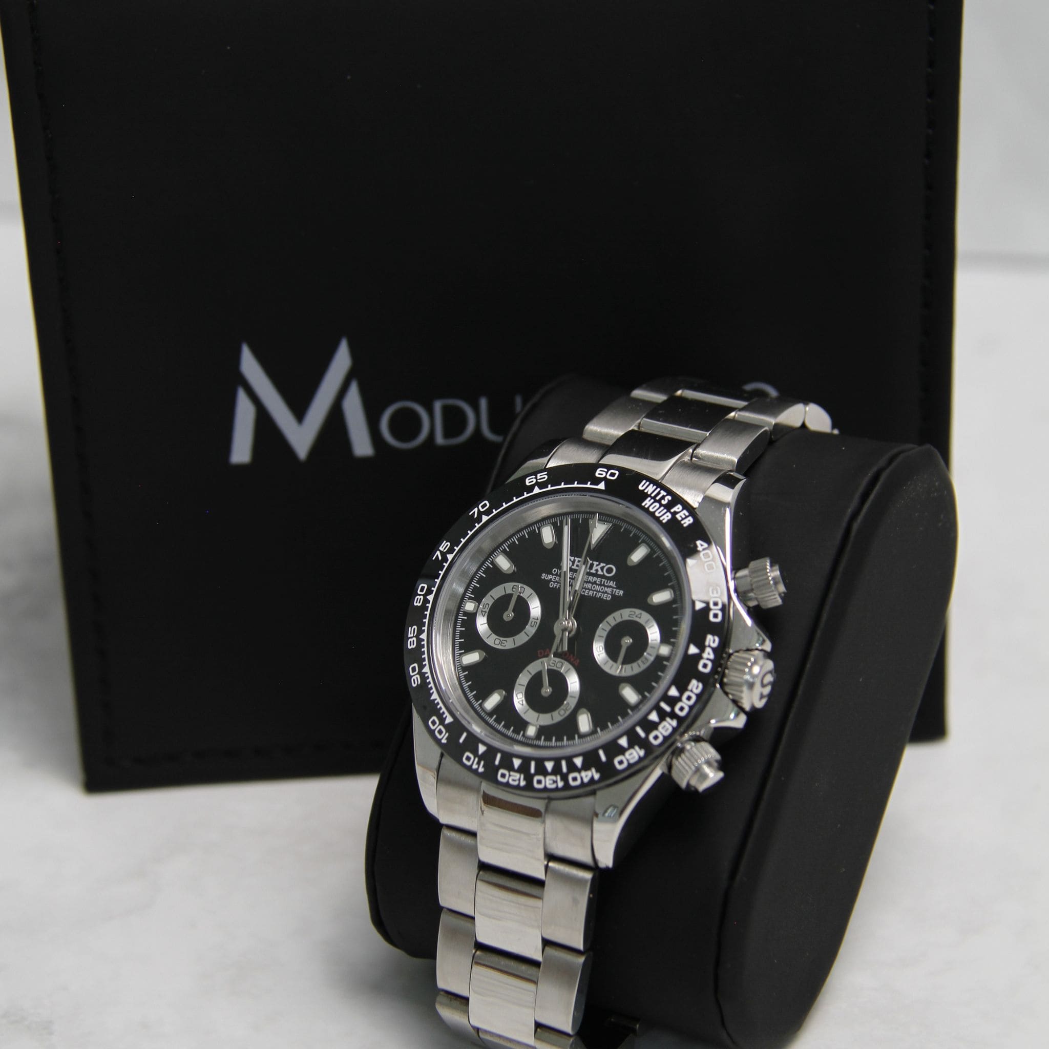 Montre Seiko Mod daytona noir sur coussin noir avec écrin en arrière plan.