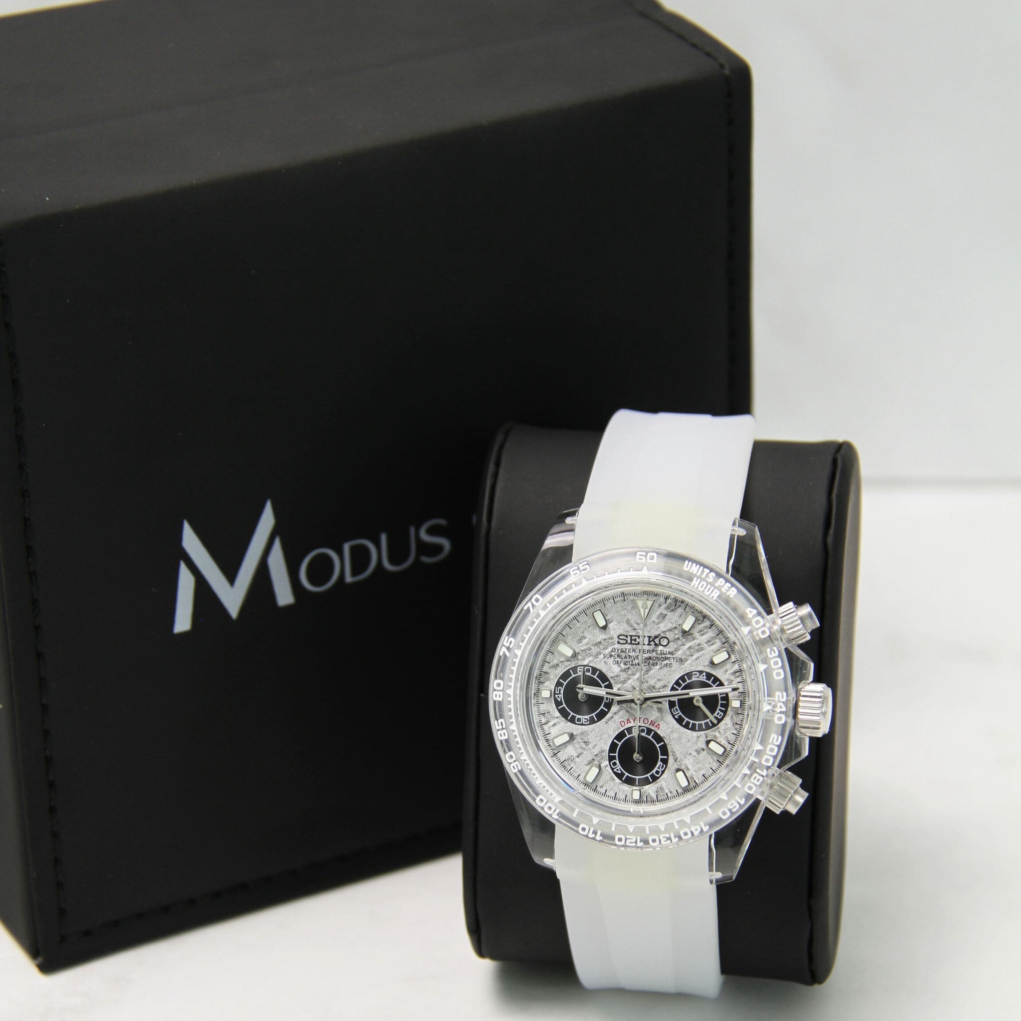 Montre Seiko Mod Daytona oysterflex gris sur coussin noir avec écrin en arrière plan.