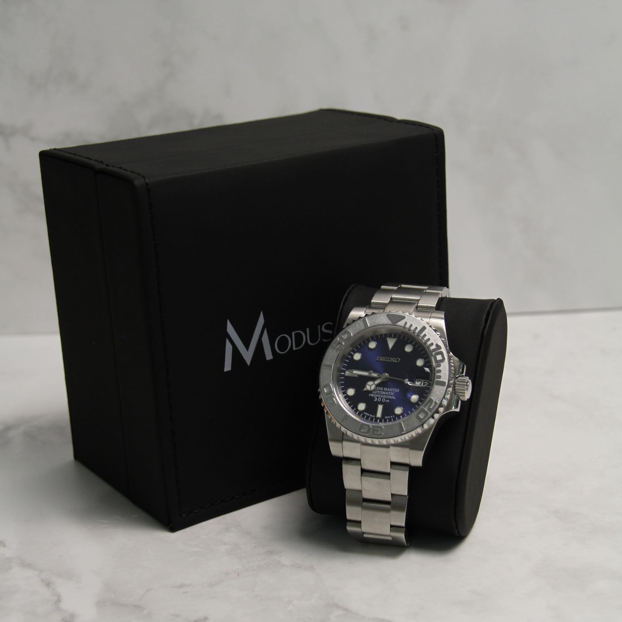 Montre Seiko Mod yachtmaster bleu sur coussin noir avec écrin en arrière plan. 
